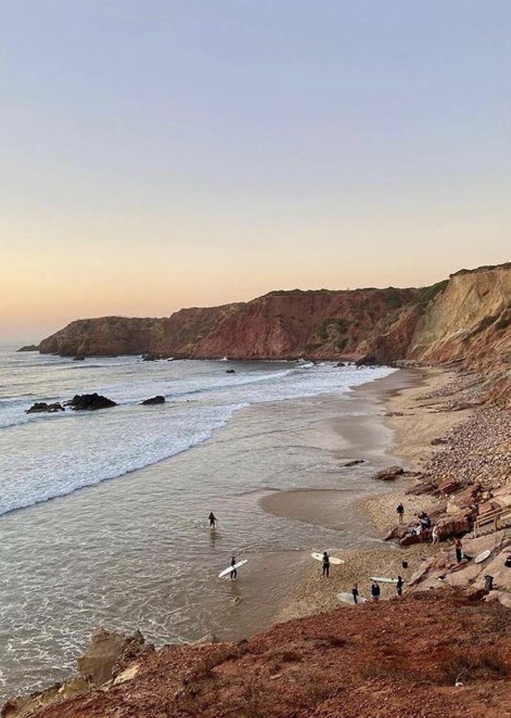 Surfers walking a wide bay at dusk beneath red cliffs, Algarve